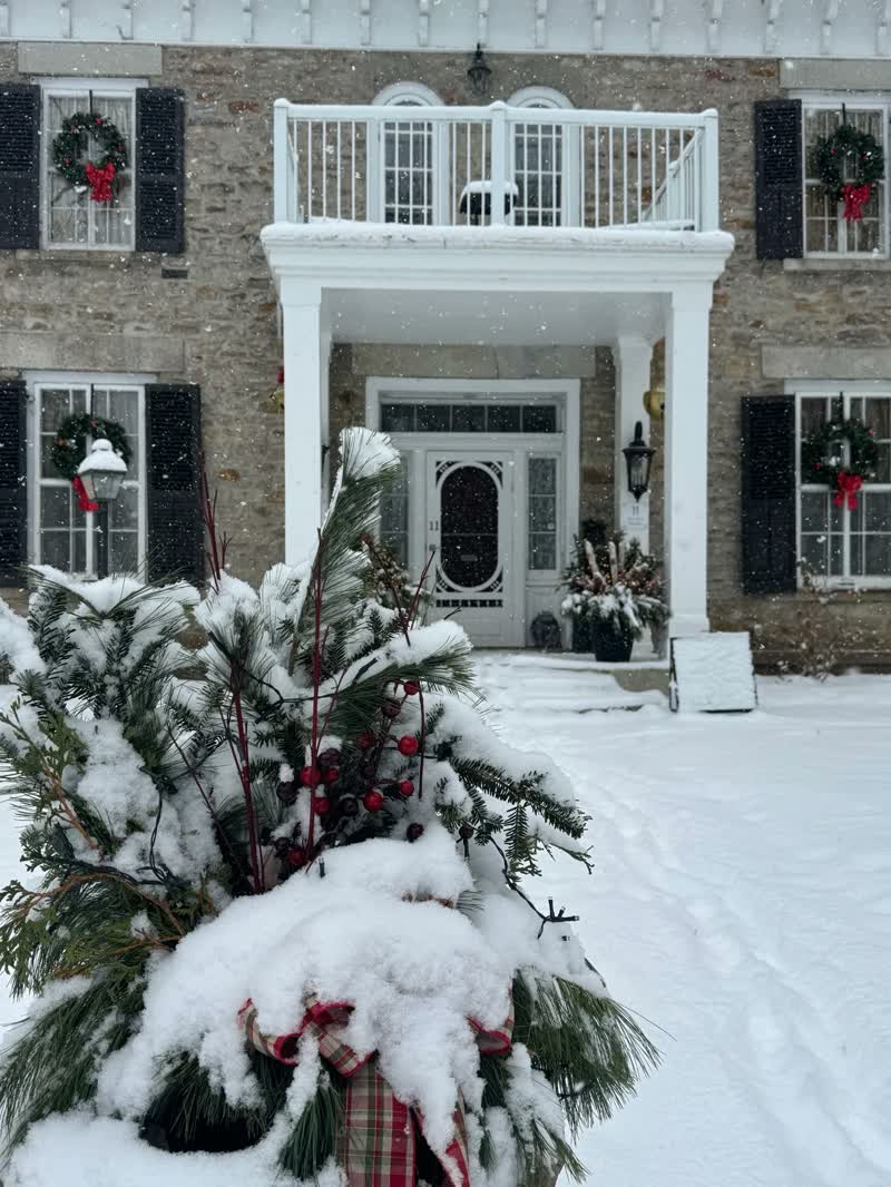 Snow falls in front of Newboro Manor’s stone façade and holiday-dressed entrance.